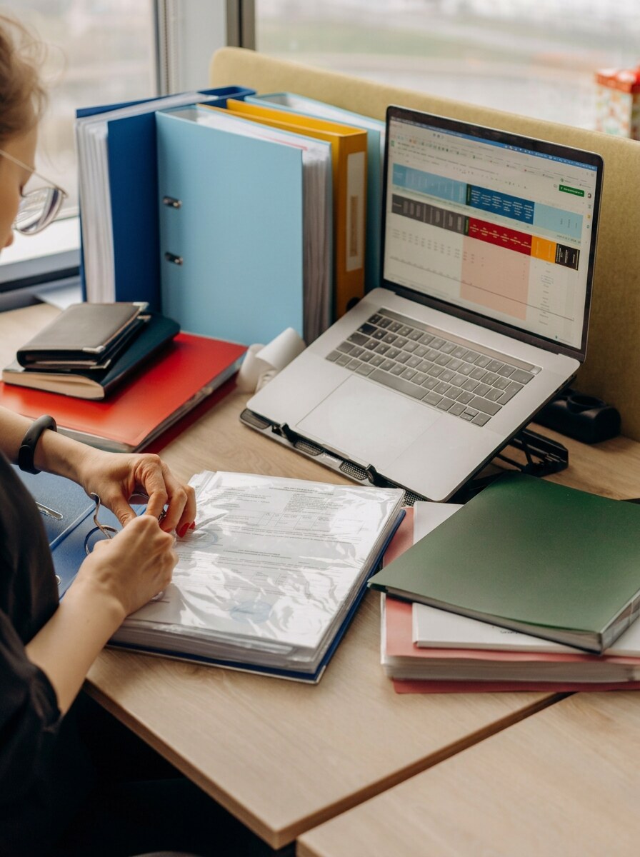 A compliance practitioner working through ring binders and a laptop at a tidy desk in warm window light &mdash; the operational reality behind an environmental approval lodgement